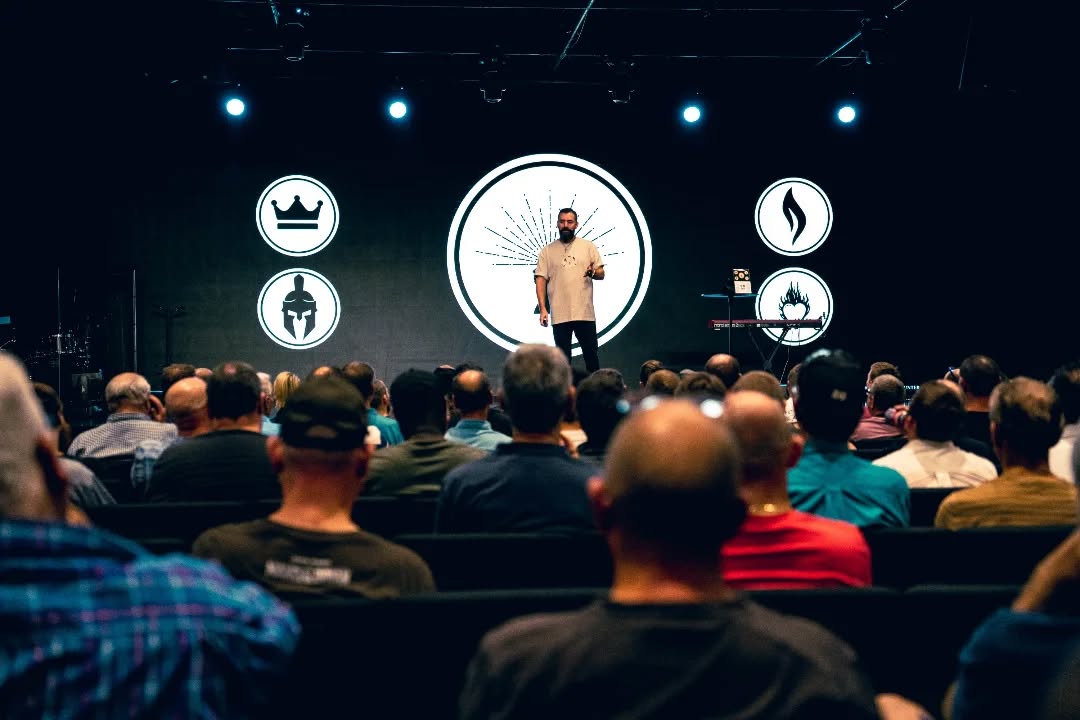 A speaker presents on stage to an audience at an event, with various icons displayed on a large screen behind them. THis is Fightmind skills and personal development training in mindset for men, women and youth
